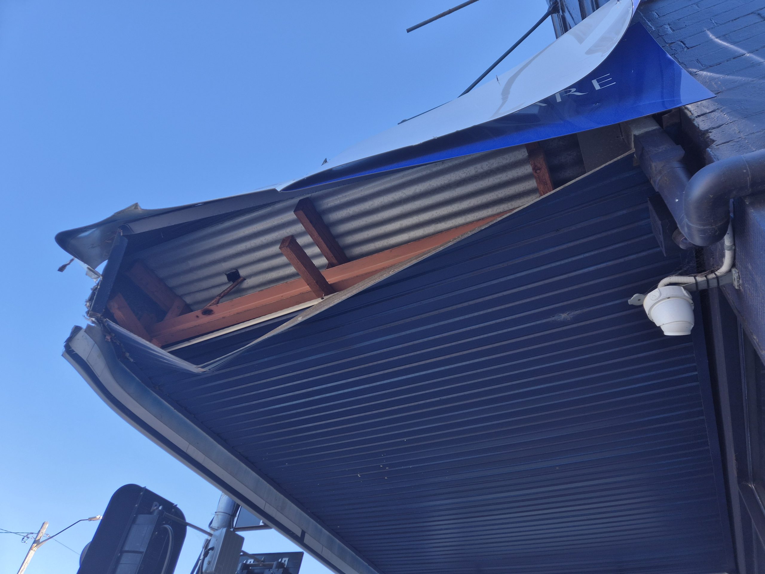 Awning collapsed onto a shopfront after a vehicle impact, with workers clearing debris behind hazard tape