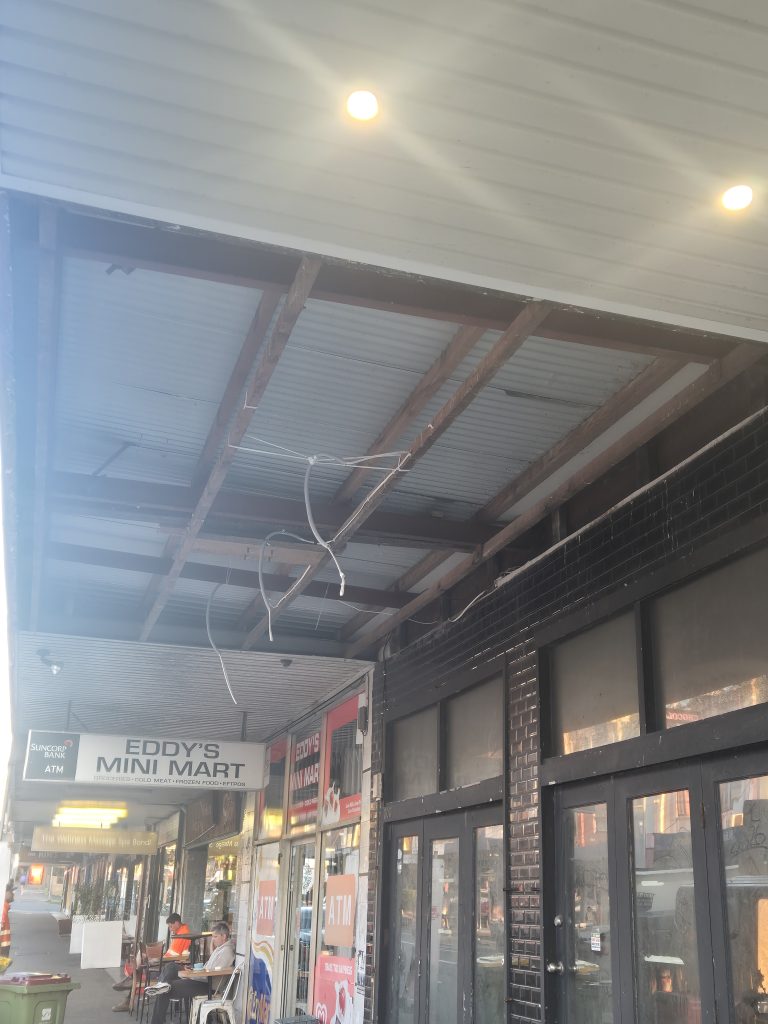 Underside of a Bondi Road street awning stripped back to the steel frame and roof sheeting, with pedestrians and outdoor seating on the footpath below